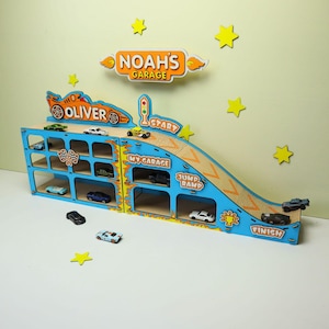 May include: A blue wooden toy car garage and ramp display with multiple parking slots. It features orange flame accents, yellow stars, and text including "NOAH'S GARAGE," "OLIVER," "START," "MY GARAGE," "JUMP RAMP," and "FINISH." Several small toy cars are included.