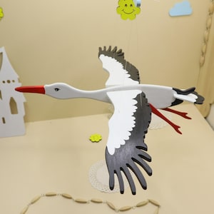 Op de afbeelding: Een houten mobiel van een vliegende ooievaar, geschilderd in wit, grijs en zwart, met een rode snavel en poten. De vleugels zijn wijd gespreid en de mobiel hangt aan een dun touwtje. Decoratieve elementen: gele bloem en wit kasteel.