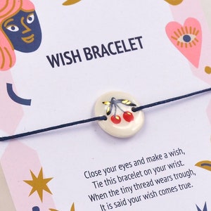 Peut inclure: Une carte rose avec un bracelet à vœux attaché. La carte présente un motif coloré avec des étoiles, des cœurs et un croissant de lune. Le bracelet a un charme en céramique blanche avec un motif de cerise rouge. La carte indique "WISH BRACELET" et comprend des instructions sur la façon de faire un vœu.
