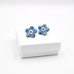 Puede incluir: Un par de pendientes de botón de flores azules y doradas. Las flores tienen centros blancos y detalles dorados.