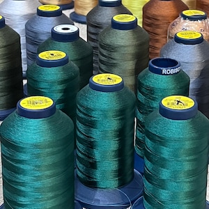 May include: A collection of spools of thread in various colors, including green, brown, gray, and black. The spools are arranged in a circular pattern on a table. The spools are labeled with the brand name "Robison-Anton" and the text "Made in USA".