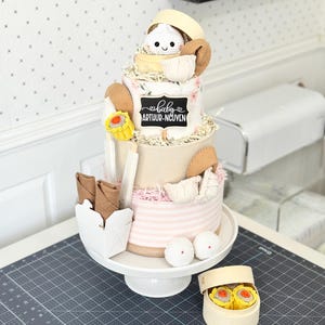 May include: A multi-tiered baby shower cake with a dim sum theme. The cake features a white crocheted baby, various dim sum items, and the name "ARTHUR-NGUYEN". The cake is displayed on a white cake stand.