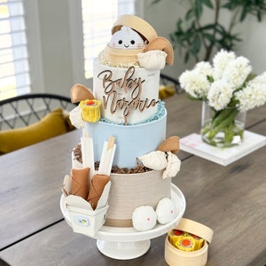 May include: A three-tiered diaper cake decorated with a white crocheted dumpling, brown chopsticks, and a white takeout box with two brown spring rolls. The cake is decorated with a wooden sign that says "Baby Mazurie".