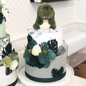 Peut inclure: Un g&acirc;teau de couches &agrave; deux &eacute;tages d&eacute;cor&eacute; d'une peluche dinosaure verte, de feuilles de monstera en feutrine et de formes succulentes. La base est envelopp&eacute;e de tissu gris avec un ruban vert fonc&eacute; et un &oelig;uf cr&egrave;me tachet&eacute; nich&eacute; dans le feuillage.