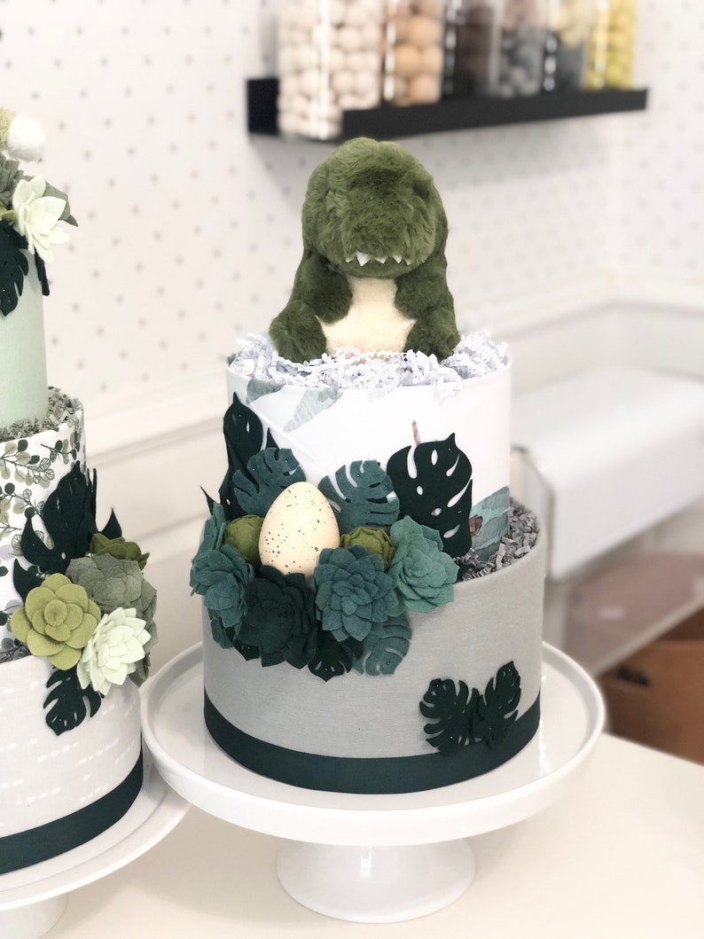 Peut inclure: Un g&acirc;teau de couches &agrave; deux &eacute;tages d&eacute;cor&eacute; d'une peluche dinosaure verte, de feuilles de monstera en feutrine et de formes succulentes. La base est envelopp&eacute;e de tissu gris avec un ruban vert fonc&eacute; et un &oelig;uf cr&egrave;me tachet&eacute; nich&eacute; dans le feuillage.