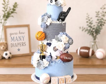 May include: A four-tier diaper cake decorated with a sports theme, featuring a soccer ball, basketball, football, and baseball. It includes a gold trophy, a black foam finger, white and blue fabric flowers, and wooden blocks spelling out MVP.