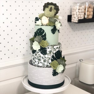 Peut inclure: Un g&acirc;teau d&eacute;coratif &agrave; trois &eacute;tages avec un niveau sup&eacute;rieur vert clair, un niveau central &agrave; motif de feuilles vertes et un niveau inf&eacute;rieur gris avec des tirets blancs. Il est orn&eacute; de succulentes en feutrine, de feuilles de monstera et d'un tric&eacute;ratops.
