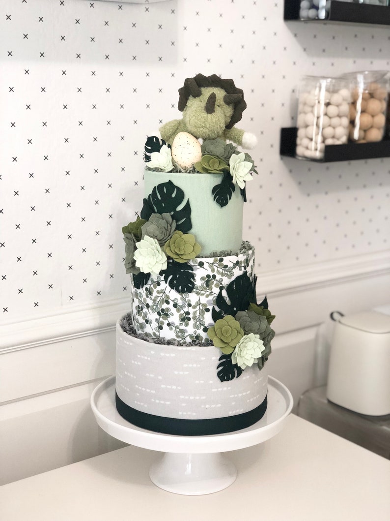 Peut inclure: Un g&acirc;teau d&eacute;coratif &agrave; trois &eacute;tages avec un niveau sup&eacute;rieur vert clair, un niveau central &agrave; motif de feuilles vertes et un niveau inf&eacute;rieur gris avec des tirets blancs. Il est orn&eacute; de succulentes en feutrine, de feuilles de monstera et d'un tric&eacute;ratops.