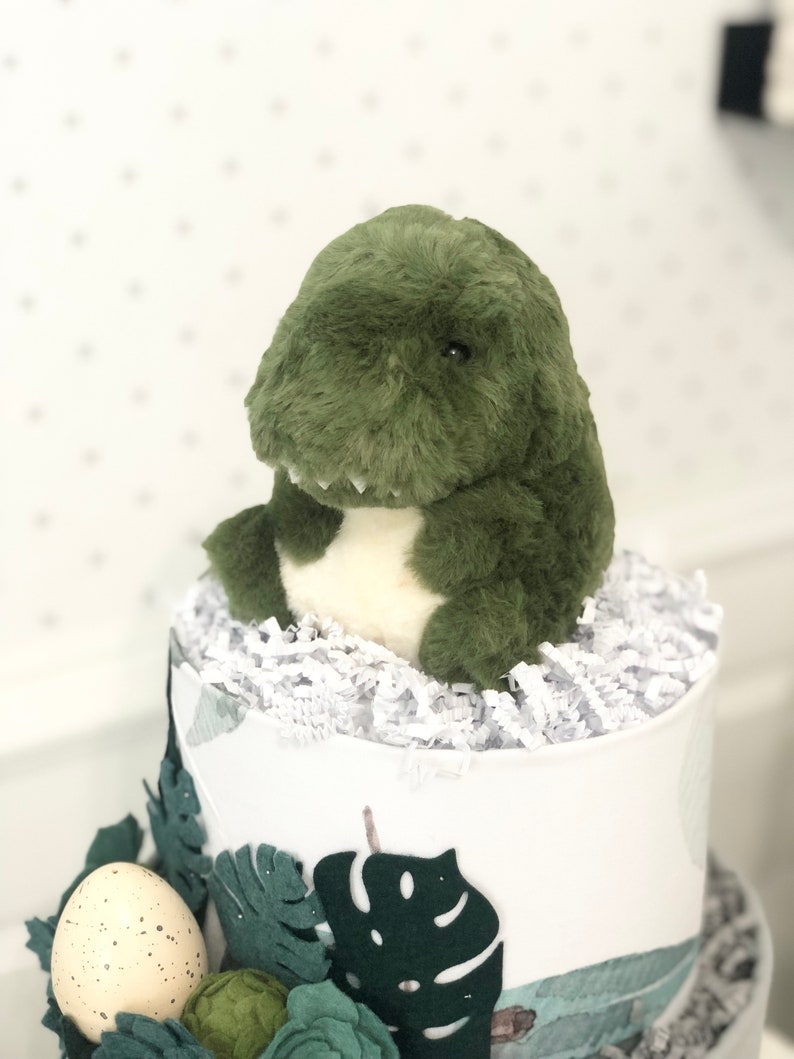 Peut inclure: Une peluche dinosaure verte avec un ventre cr&egrave;me est pos&eacute;e sur un contenant cadeau cylindrique rempli de papier d&eacute;chiquet&eacute; blanc. Le contenant est d&eacute;cor&eacute; de feuilles de monstera en feutrine et d'un ornement en forme d'&oelig;uf tachet&eacute;.