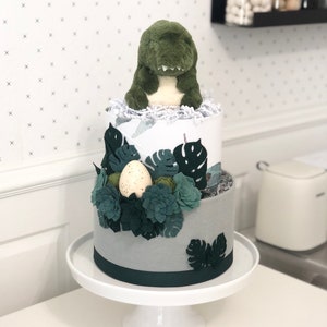 Peut inclure: Un g&acirc;teau de couches &agrave; deux &eacute;tages surmont&eacute; d'une peluche dinosaure verte, avec du papier froiss&eacute; blanc et une d&eacute;coration compos&eacute;e de feuilles de monstera en feutrine vert fonc&eacute;, de plantes grasses et d'un &oelig;uf cr&egrave;me tachet&eacute; sur un socle blanc.