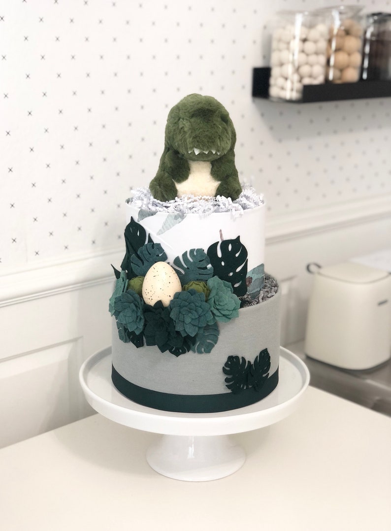 Peut inclure: Un g&acirc;teau de couches &agrave; deux &eacute;tages surmont&eacute; d'une peluche dinosaure verte, avec du papier froiss&eacute; blanc et une d&eacute;coration compos&eacute;e de feuilles de monstera en feutrine vert fonc&eacute;, de plantes grasses et d'un &oelig;uf cr&egrave;me tachet&eacute; sur un socle blanc.