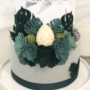 Peut inclure: Un g&acirc;teau cylindrique &agrave; deux &eacute;tages d&eacute;cor&eacute; de tissu blanc et gris, orn&eacute; de feuilles de monstera en feutrine vert fonc&eacute;, de diverses succulentes en feutrine verte et d'un &oelig;uf cr&egrave;me tachet&eacute; de brun, surmont&eacute; d'une cr&eacute;ature en peluche verte.