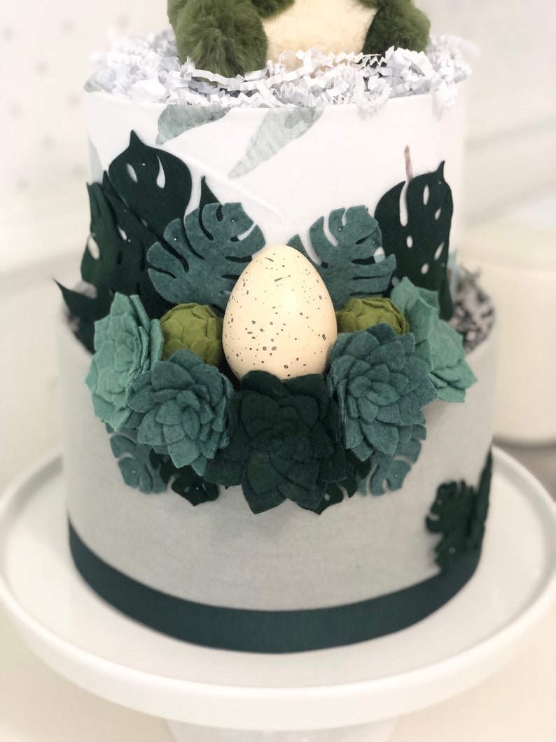 Peut inclure: Un g&acirc;teau cylindrique &agrave; deux &eacute;tages d&eacute;cor&eacute; de tissu blanc et gris, orn&eacute; de feuilles de monstera en feutrine vert fonc&eacute;, de diverses succulentes en feutrine verte et d'un &oelig;uf cr&egrave;me tachet&eacute; de brun, surmont&eacute; d'une cr&eacute;ature en peluche verte.