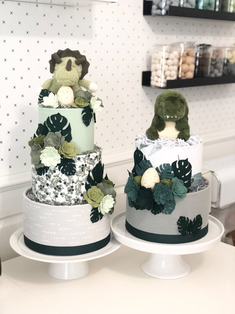 Peut inclure: Deux g&acirc;teaux de couches d&eacute;cor&eacute;s de dinosaures en peluche, de plantes succulentes en feutrine et de feuilles de monstera. Le g&acirc;teau de gauche a trois &eacute;tages en tissu vert clair, imprim&eacute; botanique et gris. Celui de droite a deux &eacute;tages gris et blanc.