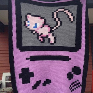 May include: A purple and black crocheted blanket featuring a pixelated image of a pink Pokemon character on a grey background. The blanket is shaped like a Game Boy.