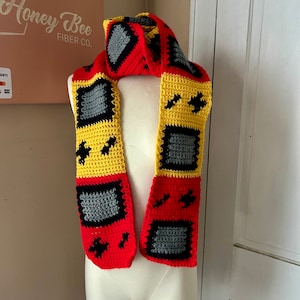 May include: A crocheted scarf in red, yellow, black, and gray, resembling a vintage handheld video game console. The scarf is displayed on a white mannequin.