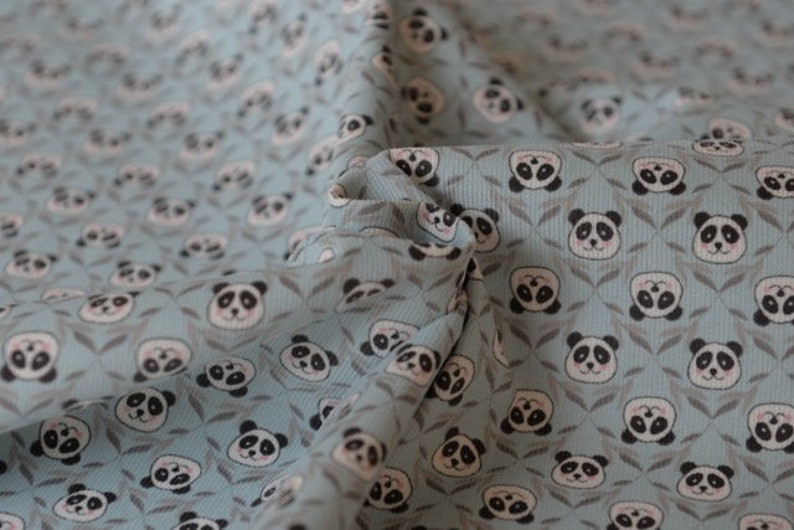 K&ouml;nnte beinhalten: Ein blauer Stoff mit einem sich wiederholenden Muster von Pandab&auml;ren-Gesichtern. Die Pandas sind wei&szlig; mit schwarzen Augen und Ohren und von kleinen gr&uuml;nen Bl&auml;ttern umgeben.
