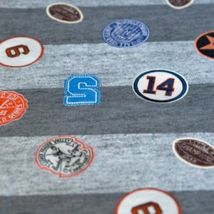 Peut inclure: Un tissu rayé gris et blanc avec divers patchs brodés. Les patchs comprennent un logo d'équipe de baseball, une étoile, le numéro "14" et les lettres "S" et "6".