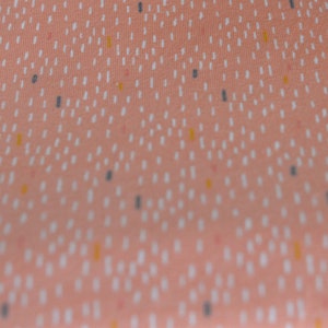 May include: A close-up of a peach-colored fabric with a repeating pattern of white, yellow, and gray dashes.