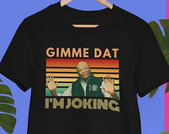 May include: Black t-shirt with the text "GIMME DAT I'M JOKING" in a retro style. The design features a man with his arms outstretched. The shirt is hanging on a wooden hanger.
