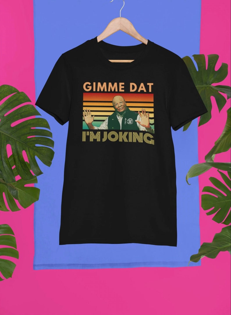 May include: Black t-shirt with the text "GIMME DAT I'M JOKING" in a retro style. The design features a man with his arms outstretched. The shirt is hanging on a wooden hanger.