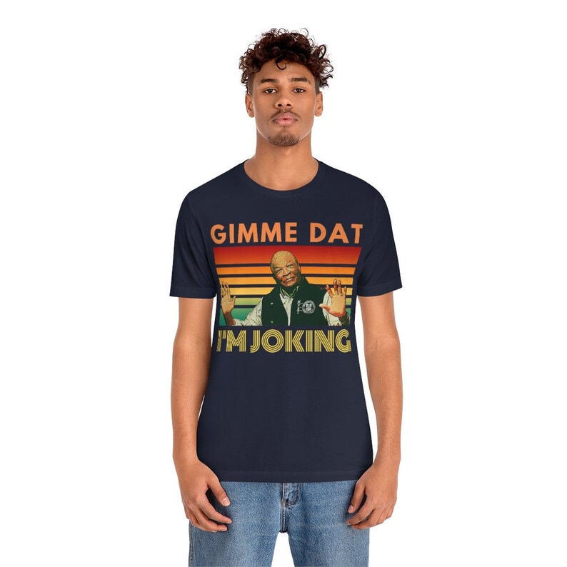 May include: Navy blue t-shirt featuring a retro design with the text "GIMME DAT I'M JOKING". The graphic includes an image of a man with his hands up. The shirt is a unisex style.
