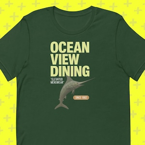 May include: Dark green t-shirt with the text "OCEAN VIEW DINING" in a light yellow font. Below the text is a graphic of a marlin fish. The text "ELEVATED MENSWEAR" and "SINCE 1982" are also printed on the shirt.