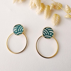 Op de afbeelding: Een paar goudkleurige oorringen. Elke oorring heeft een cirkelvormige, blauwgroene en witte patroon top. De oorbellen worden gepresenteerd op een witte ondergrond, met gedroogde beige bloemen op de achtergrond. Een modeaccessoire.
