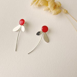 Peut inclure: Une paire de boucles d'oreilles en argent avec des accents de fleurs rouges. Chaque boucle d'oreille présente un cercle rouge unique avec trois pétales argentés et une longue tige argentée fine.