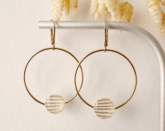 May include: A pair of gold-colored hoop earrings. Each earring features a clear, round bead with horizontal gold stripes. The earrings are suspended from a silver bar, with dried flowers in the background. The earrings are a simple, elegant design.