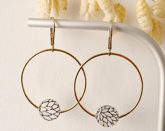 May include: A pair of gold hoop earrings with a small, round, white and black patterned bead at the bottom of each hoop. The earrings are displayed on a silver metal stand with dried, light-colored flowers in the background.