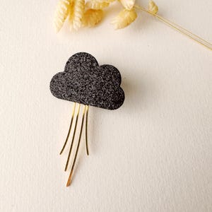 以下が含まれることがあります： 金色メタルのアクセントが付いた黒い雲の形をしたブローチ。雲はキラキラした質感で、金色のアクセントは細くてまっすぐです。
