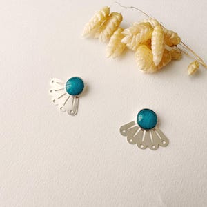 May include: A pair of silver-toned fan-shaped earrings with a round, turquoise-colored stone in the center. The earrings are displayed on a white surface, with dried beige flowers in the background.