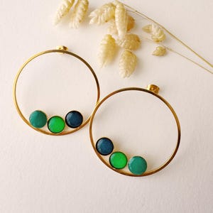 Op de afbeelding: Een paar goudkleurige oorringen. Elke oorring heeft drie groene en blauwe cirkelvormige accenten. De oorbellen worden gepresenteerd op een wit oppervlak met gedroogde, beige plantenstengels op de achtergrond.