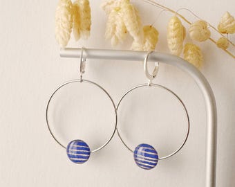 May include: A pair of silver hoop earrings with a blue and white striped bead at the bottom of each hoop. The earrings are displayed on a silver stand with dried, beige-colored flowers.