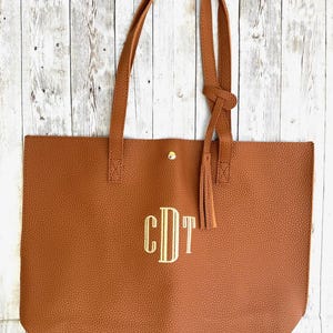 Op de afbeelding: Een bruine leren tote bag met een gouden kwast en een gouden knoopsluiting. De tas heeft een monogram van de letters "CDT" in gouden draad.