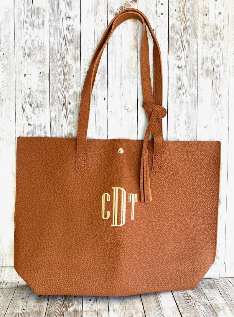Op de afbeelding: Een bruine leren tote bag met een gouden kwast en een gouden knoopsluiting. De tas heeft een monogram van de letters "CDT" in gouden draad.