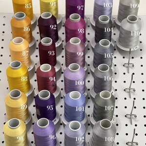Op de afbeelding: Een wanddisplay met 18 spoelen garen in verschillende kleuren, waaronder geel, paars, grijs en blauw. De spoelen zijn genummerd van 85 tot en met 110.