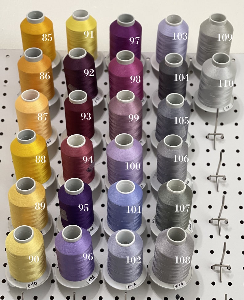 Op de afbeelding: Een wanddisplay met 18 spoelen garen in verschillende kleuren, waaronder geel, paars, grijs en blauw. De spoelen zijn genummerd van 85 tot en met 110.