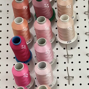 Op de afbeelding: Een verzameling van 12 spoelen garen in verschillende tinten roze en perzik. De spoelen zijn gerangschikt in rijen op een wit prikbord. Elke spoel is genummerd van 48 tot 62.