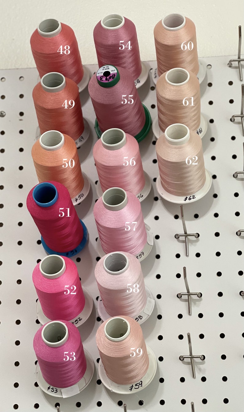 Op de afbeelding: Een verzameling van 12 spoelen garen in verschillende tinten roze en perzik. De spoelen zijn gerangschikt in rijen op een wit prikbord. Elke spoel is genummerd van 48 tot 62.