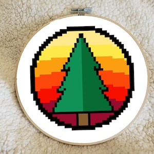 May include: A pixelated cross-stitch design of a green pine tree against a sunset gradient of yellow, orange, and red. The design is set within a white circle and framed by a wooden embroidery hoop.