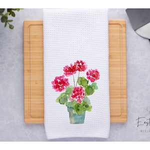 May include: White kitchen towel with a watercolor illustration of a potted pink geranium plant. The text "East Road Mercantile" is printed in the bottom right corner.