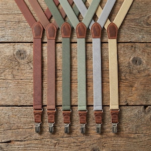 May include: A collection of six suspenders in various colors, including brown, olive green, and gray. Each suspender features leather accents and silver-toned metal clips for attachment. The suspenders are arranged on a rustic wooden surface.