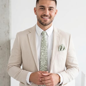 Könnte beinhalten: Ein beigefarbener Blazer über einem weißen Hemd mit Button-Down-Kragen, kombiniert mit einer salbeigrünen Krawatte und einem passenden Einstecktuch, beide mit zartem weißem Blumenmuster. Das Outfit wird durch eine goldfarbene Armbanduhr ergänzt.