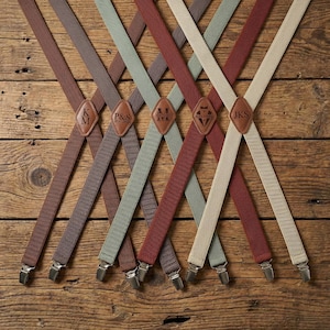 Puede incluir: Surtido de tirantes en varios colores, incluyendo marrón, gris, verde y beige, dispuestos sobre una superficie de madera. Cada tirante presenta un parche de cuero con iniciales o un diseño. Los tirantes tienen clips plateados.