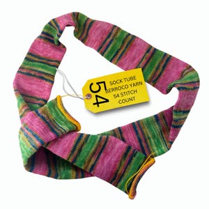 May include: A knitted sock tube in pink, green, and blue stripes with yellow trim. A yellow tag reads "SOCK TUBE BERROCO YARN 54 STITCH COUNT". The sock tube is made of yarn and is displayed against a white background.