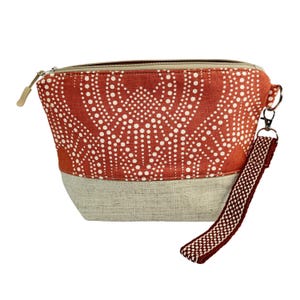 May include: A zippered pouch with a rust-colored top section featuring a white dotted pattern. The bottom section is a light beige fabric. A matching patterned wrist strap and a beige zipper complete the design. The pouch is designed for carrying small items.
