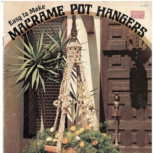 May include: A macrame plant hanger made with natural twine, hanging from a wooden door frame. The hanger is decorated with a woven design and has a brown clay pot with green plants inside. The image is from a vintage craft book titled "Easy to Make Macrame Pot Hangers".