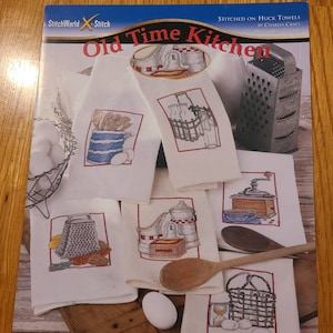 May include: A collection of white kitchen towels with cross-stitch designs featuring kitchen-themed images. The designs include a grater, a coffee grinder, and a basket of eggs. The towels are displayed on a wooden surface.